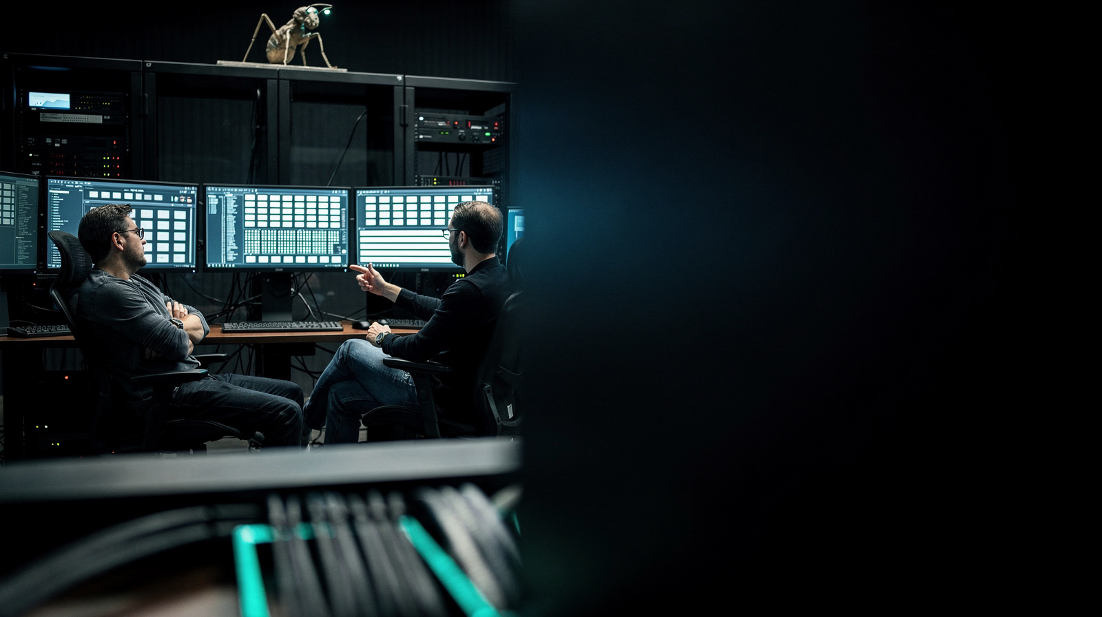 A dim control room with a wall of monitors showing real running dashboards, two people reviewing the readouts, a small mechanical ant figure on the server rack, cyan cable lighting.